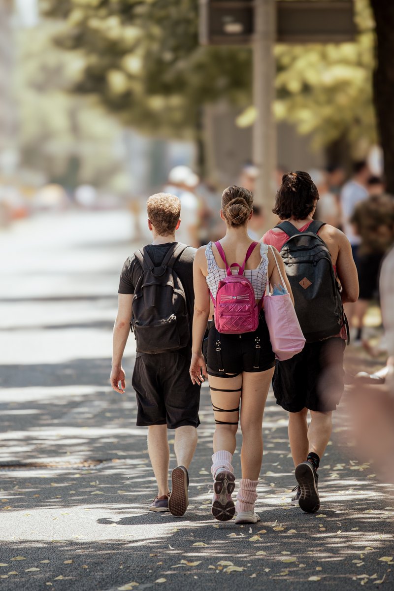Drei Besucherinnen und Besucher laufen bei sommerlicher Hitze auf dem Weg zum CSD München Pride 2026 durch eine schattige Straße