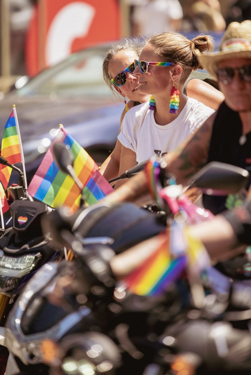 Zwei Teilnehmende mit Regenbogen-Sonnenbrillen und bunten Ohrringen sitzen auf einem Motorrad und verfolgen die Parade.