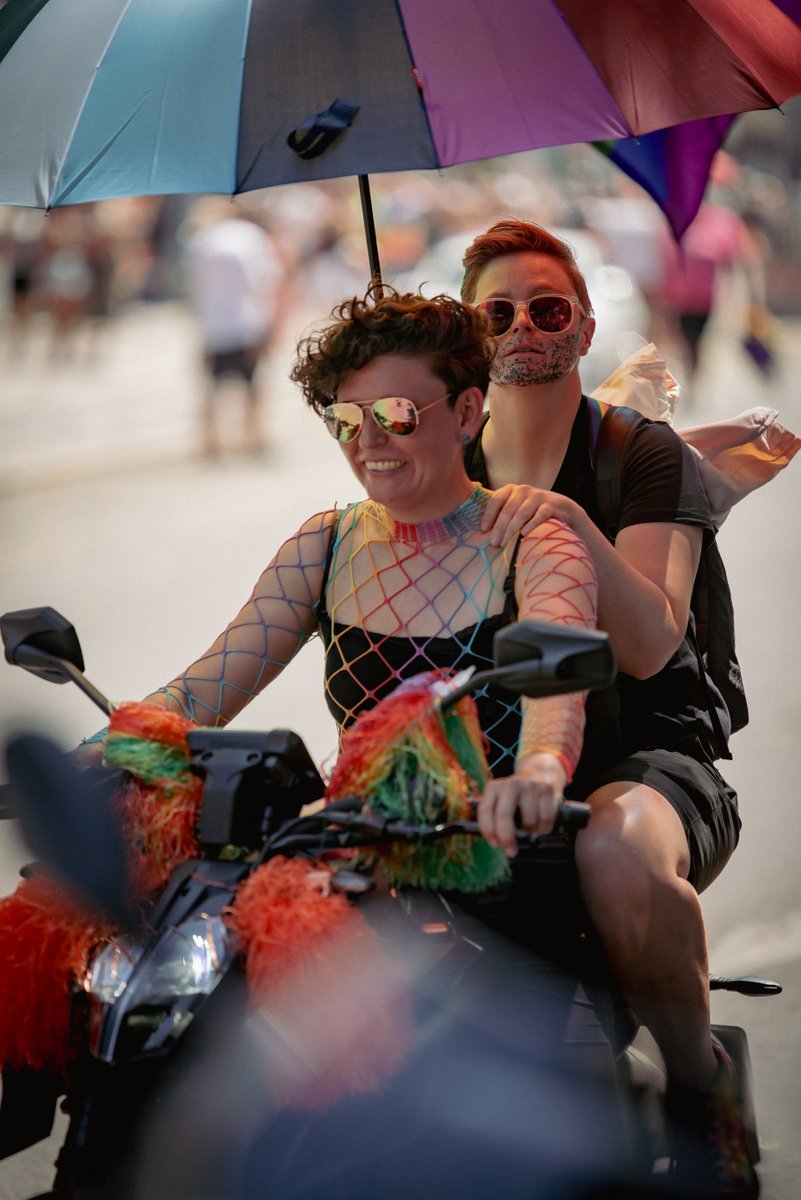 Zwei lachende Teilnehmende fahren auf einem geschmückten Roller unter einem bunten Sonnenschirm durch die Pride-Parade.