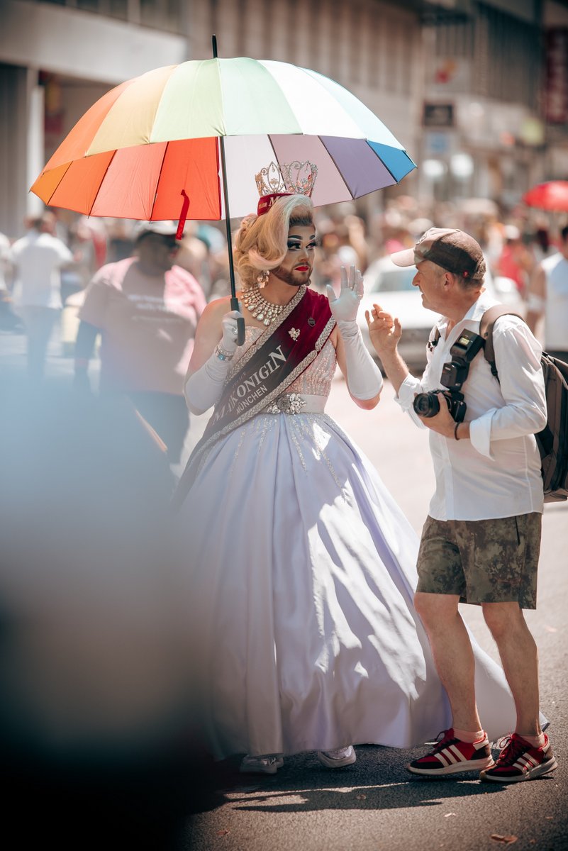 Drag Queen im eleganten weißen Kleid mit Krone und Regenbogenschirm unterhält sich mit einem Fotografen während der Parade.