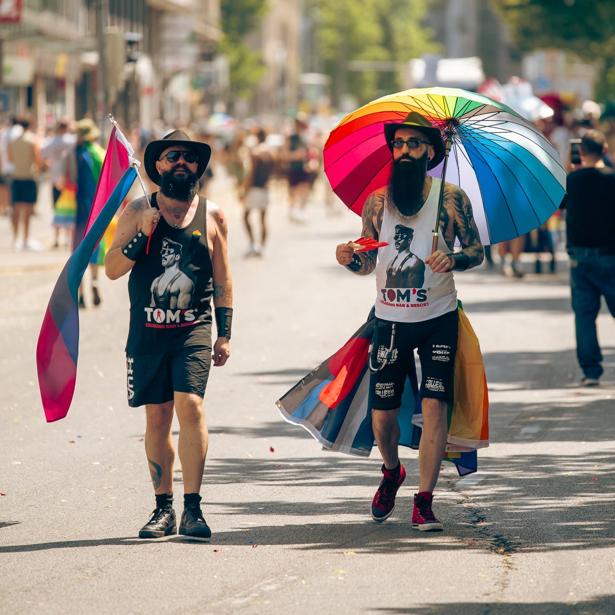 Zwei Teilnehmende in schwarzen Outfits mit Pride-Flaggen laufen unter bunten Sonnenschirmen durch die Stra?.