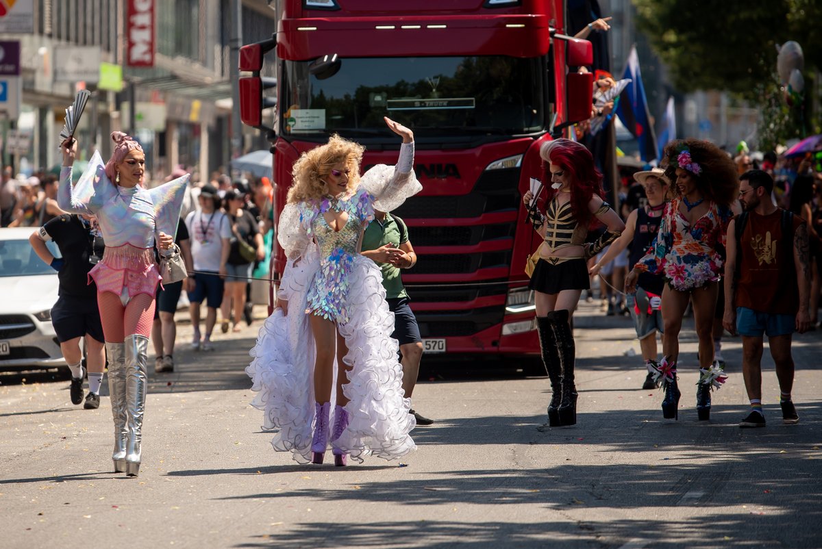Drag Queens und Teilnehmende gehen vor einem Lastwagen durch die sonnige Paradestrecke.