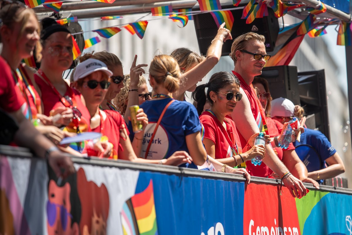 Menschen auf einem Paradewagen feiern mit Regenbogenflaggen und winken ins Publikum.